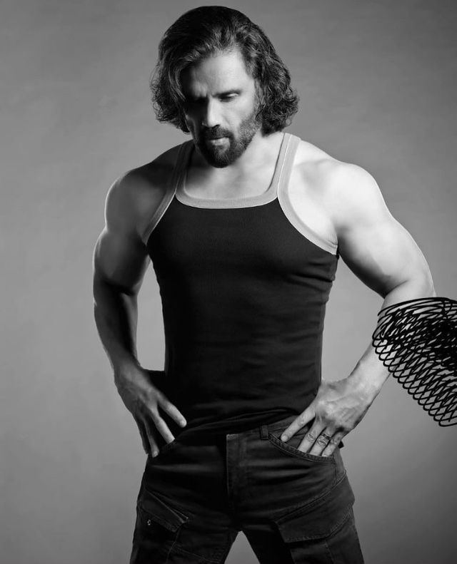 a man with his hands on his hips wearing a tank top and black jeans, posing in front of a gray background