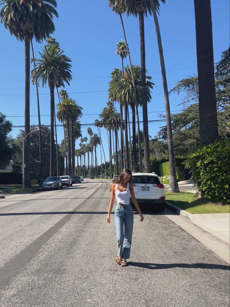 a woman is walking down the street in front of palm trees and a white car