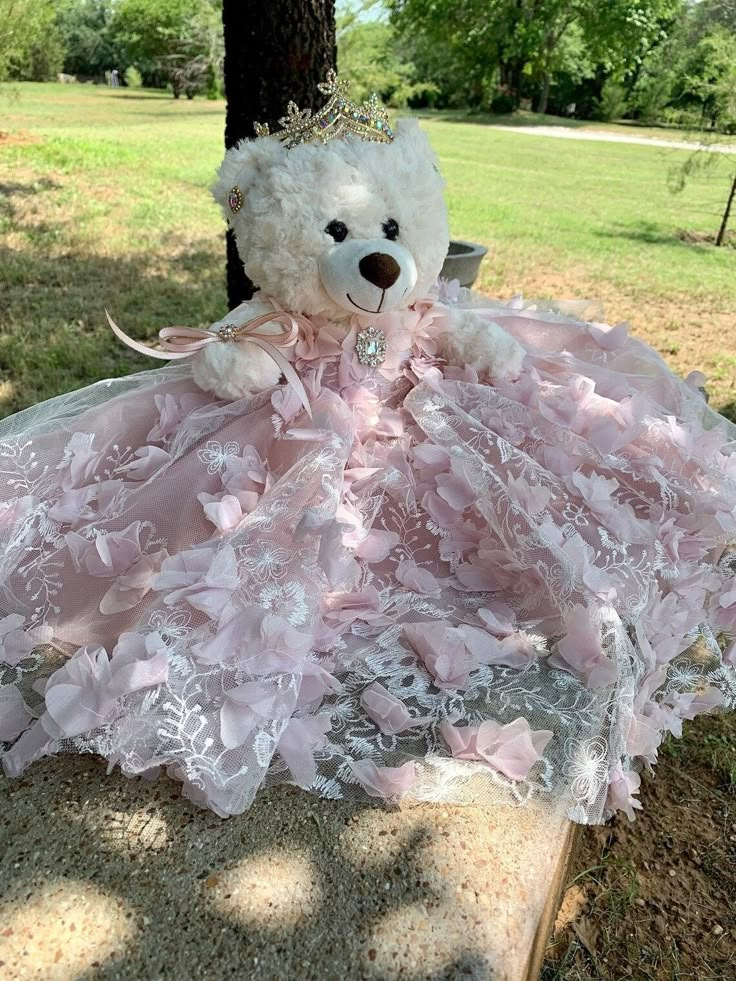 a white teddy bear wearing a pink dress sitting on a bench in the grass next to a tree