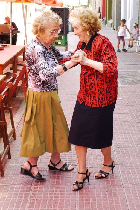 two older women standing next to each other on a brick sidewalk in front of tables and umbrellas