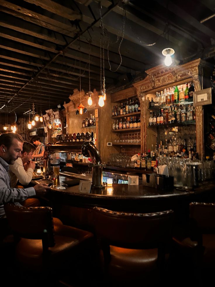 two people sitting at a bar with bottles on the shelves and lights hanging from the ceiling