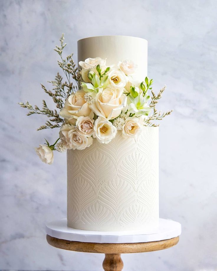 a white wedding cake decorated with flowers and greenery on a wooden stand in front of a marble background