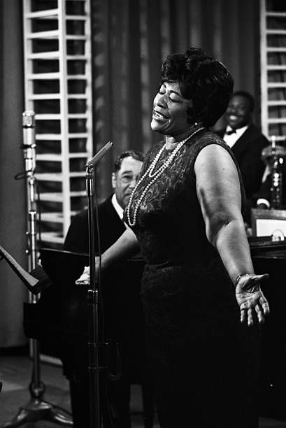 a woman standing in front of a microphone and singing into a microphone with other people behind her