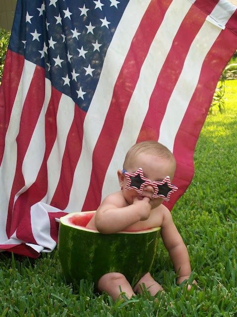 a baby sitting in a watermelon bowl on the grass with an american flag behind him