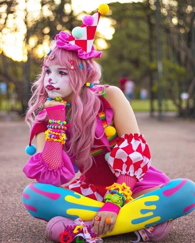 a woman with pink hair and makeup is sitting on the ground wearing colorful clothing, holding her hand up to her face