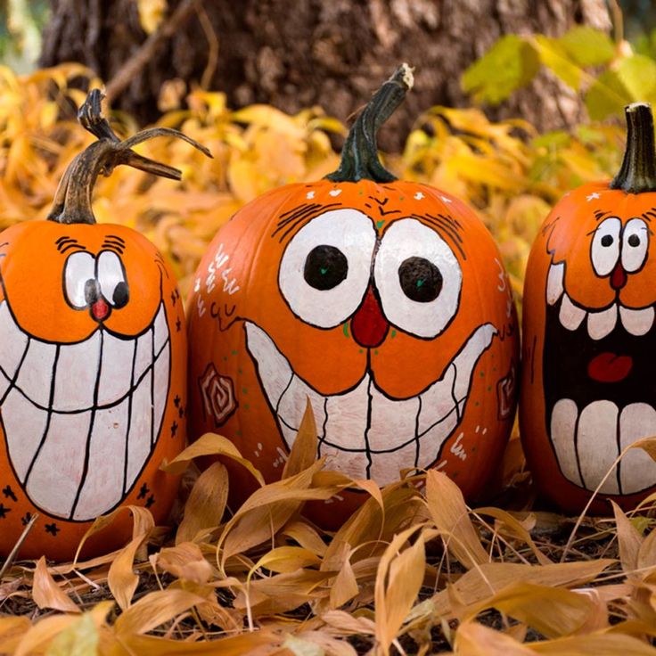 three painted pumpkins with faces on them sitting in the leaves next to a tree