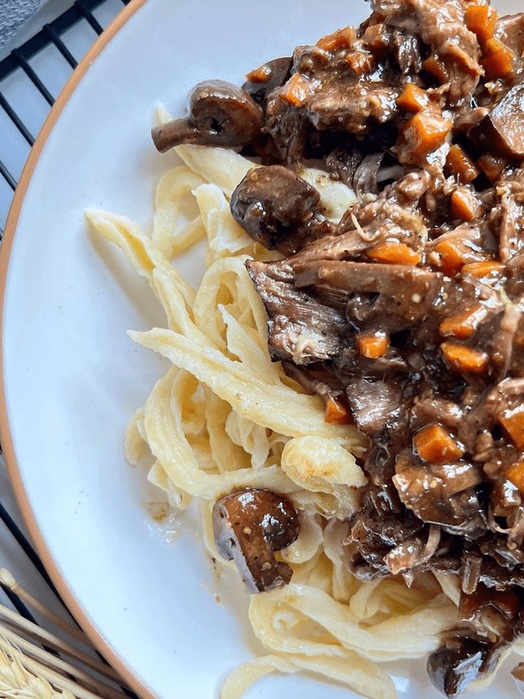 a white plate topped with pasta covered in meat and mushrooms next to a fork on top of a table