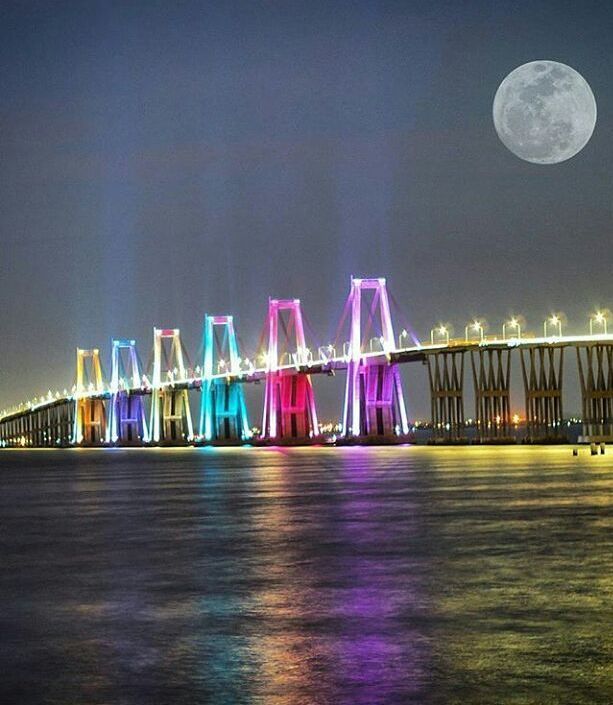 the full moon is shining brightly in the sky above a bridge over water with lights on it