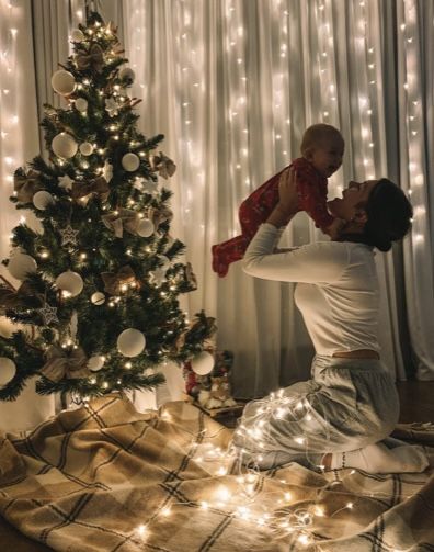 a man holding a baby in front of a christmas tree with lights on the walls