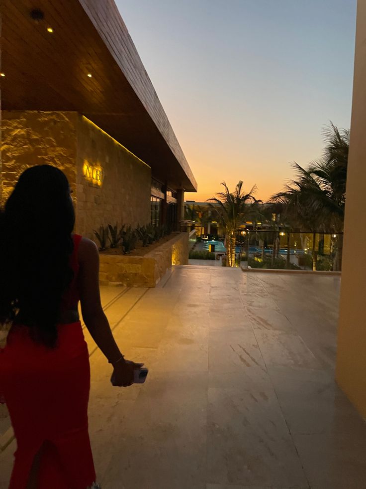 a woman in a red dress is walking towards a building with a pool and palm trees