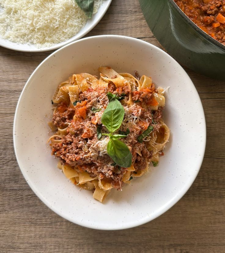 a bowl of pasta with meat and vegetables in it on a wooden table next to a pot of grated parmesan cheese