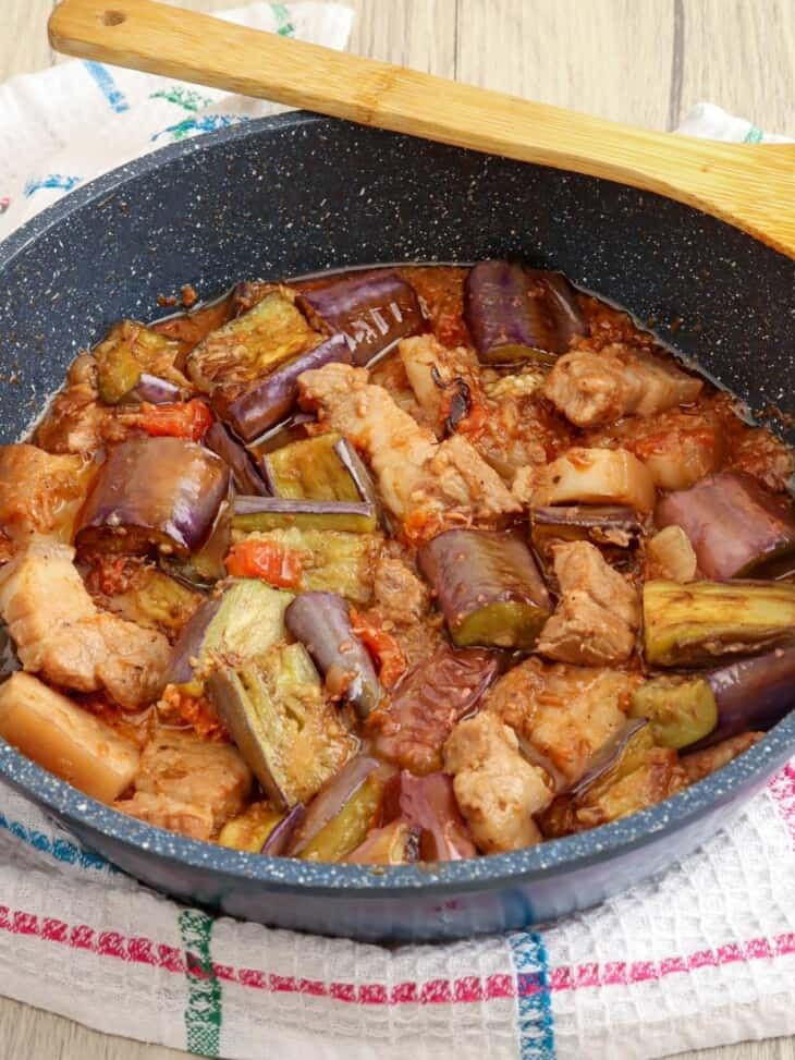 a pan filled with meat and vegetables on top of a towel next to a wooden spoon