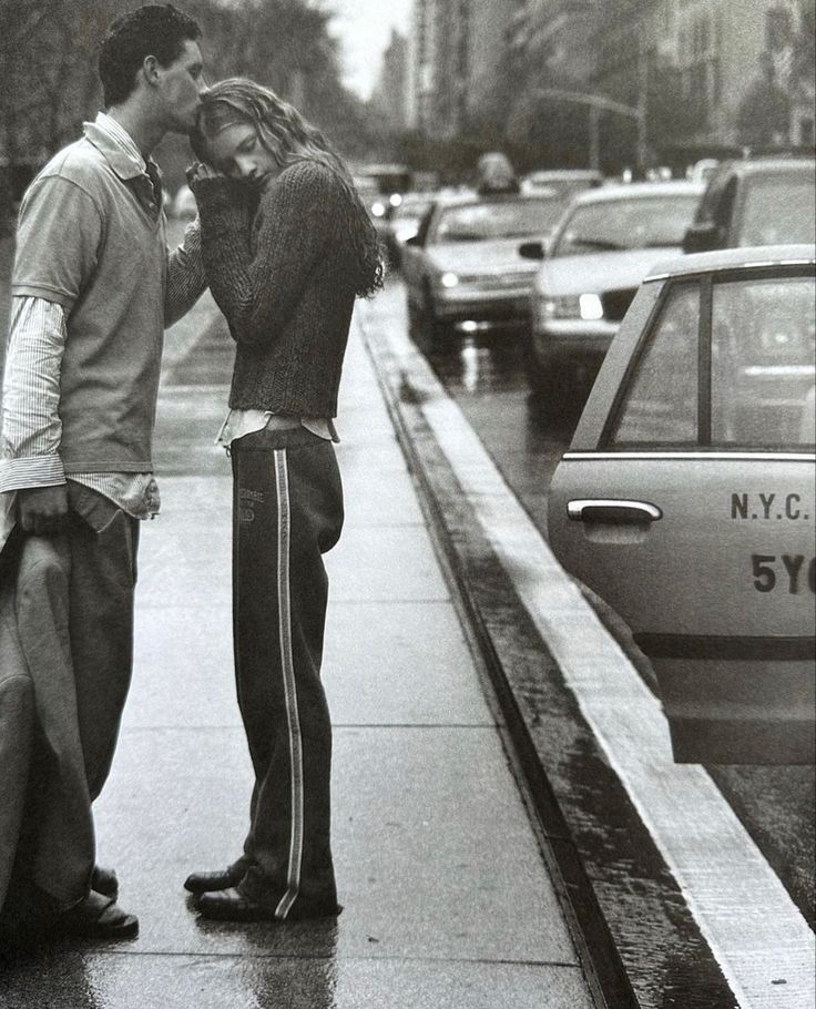 a man and woman kissing on the sidewalk in front of a parked car while it's raining