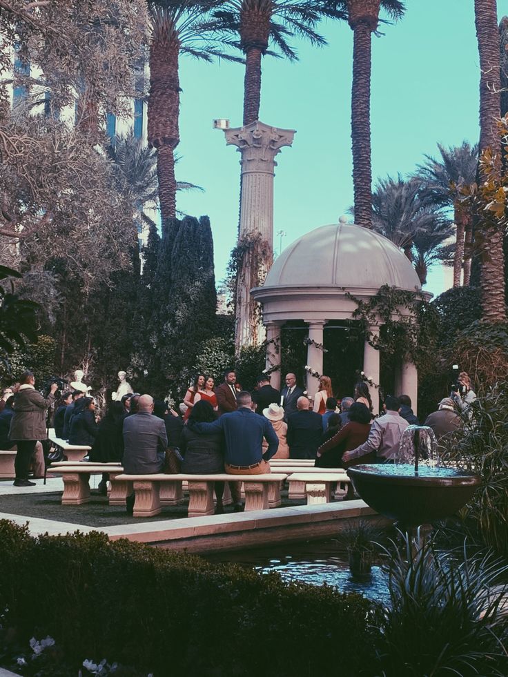 people are sitting on benches in front of a gazebo and fountain with palm trees