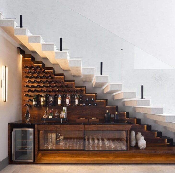 there is a wine rack under the stairs in this room, with many bottles and glasses on it