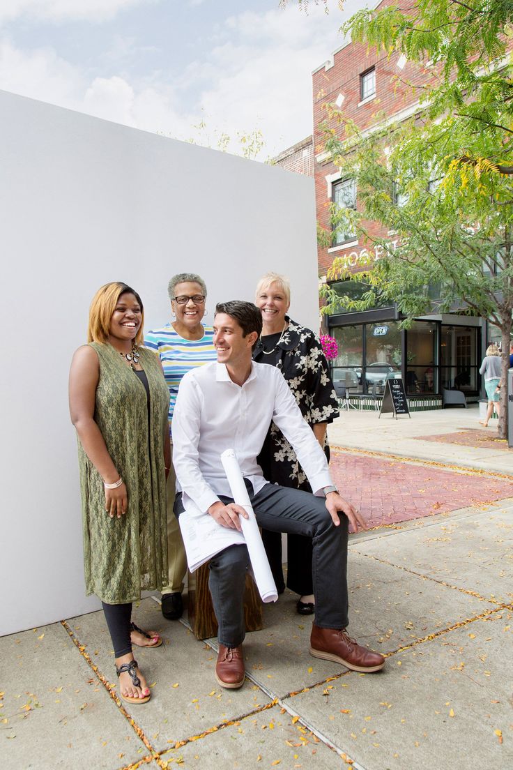 a group of people standing and sitting next to each other on a sidewalk in front of a white wall