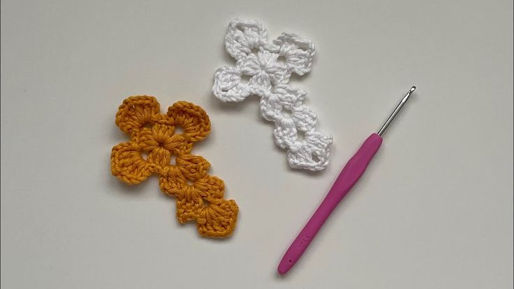 two crocheted flowers are next to a pink crochet hook on a white surface