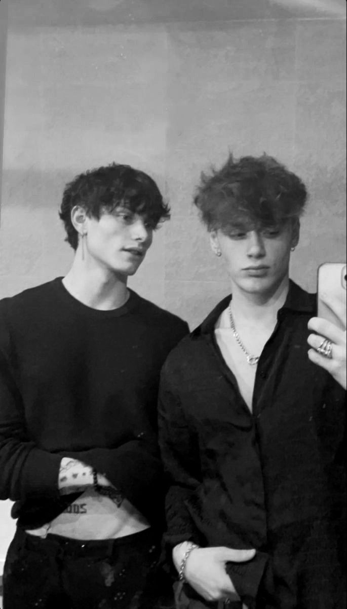 two young men standing next to each other in front of a wall holding cell phones