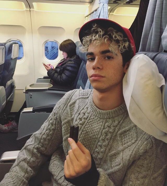 a young man sitting on an airplane holding a cell phone and looking at the camera