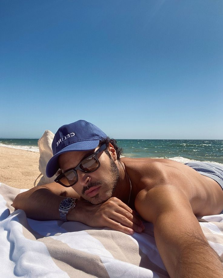 a man laying on top of a beach next to the ocean wearing a blue hat