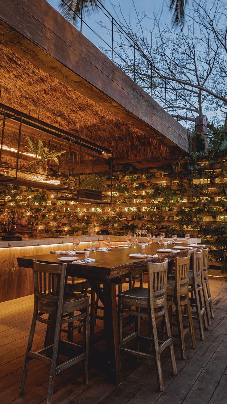 an outdoor dining area with wooden tables and chairs, surrounded by greenery at dusk