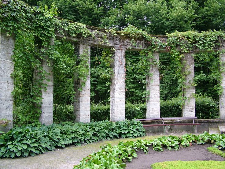 an outdoor garden with many columns and plants growing on the walls, along with a bench surrounded by greenery