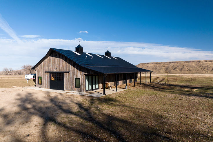 an old barn sits on the side of a dirt road in front of a field