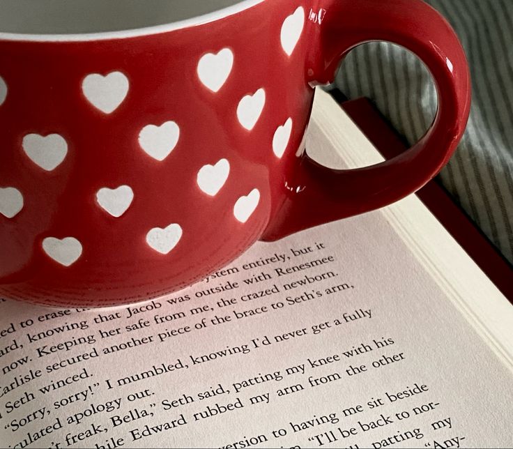 a red coffee cup sitting on top of an open book with white hearts painted on it