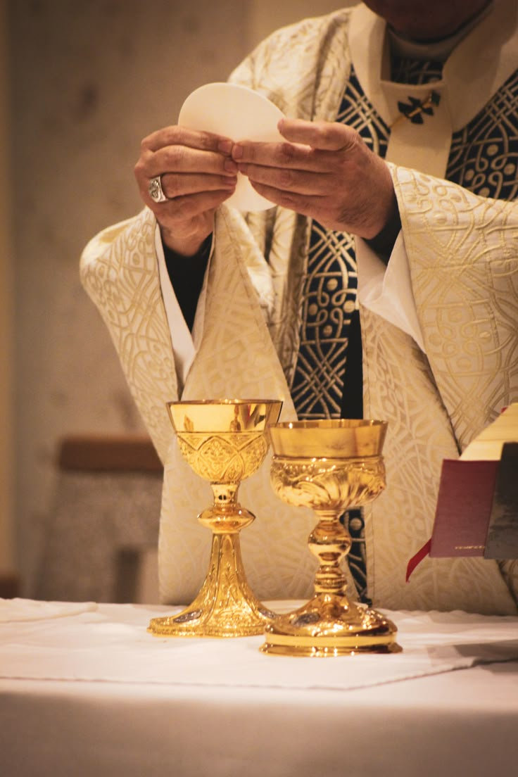 a priest is holding a white object in his hands while standing next to two golden candles