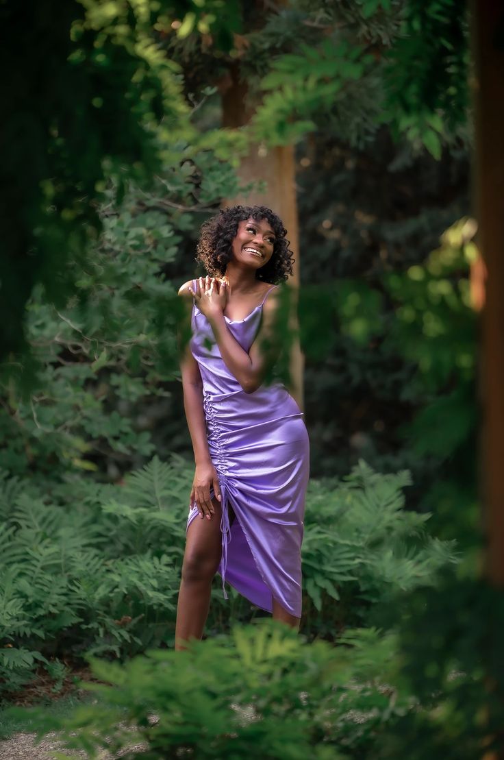 a woman in a purple dress is posing for a photo with trees and bushes behind her