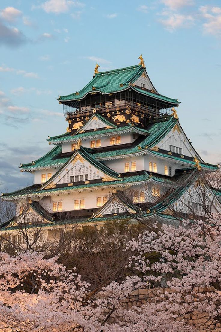 a tall white and green building surrounded by cherry blossom trees in the evening sun with clouds overhead