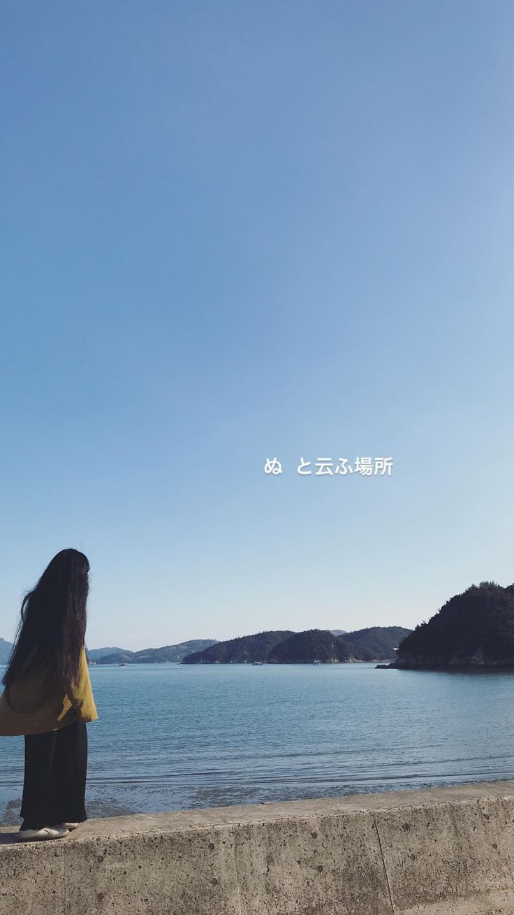 a woman sitting on top of a cement wall next to the ocean with her back turned