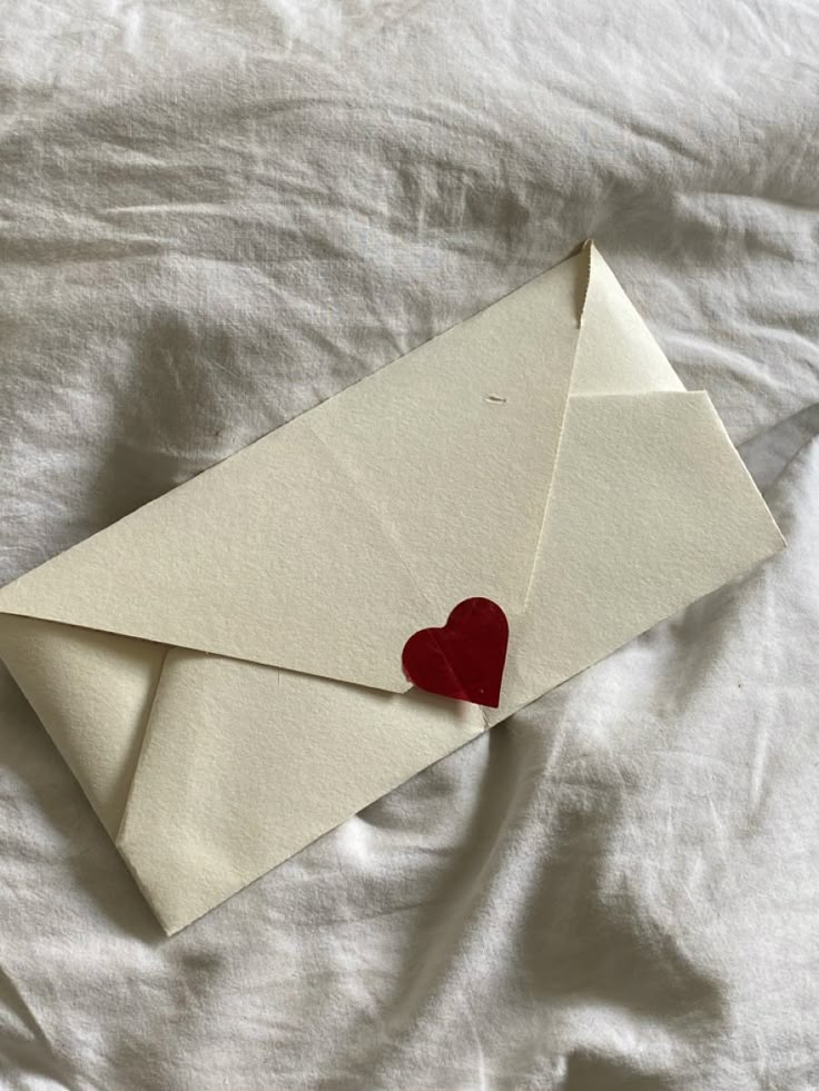 an envelope with a red heart sticking out of it on top of a white sheet