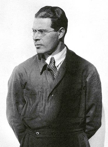 black and white photograph of a man with glasses wearing a coat, tie and dress pants