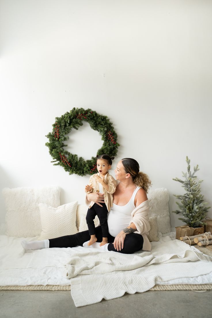 a woman sitting on top of a bed holding a baby next to a christmas wreath