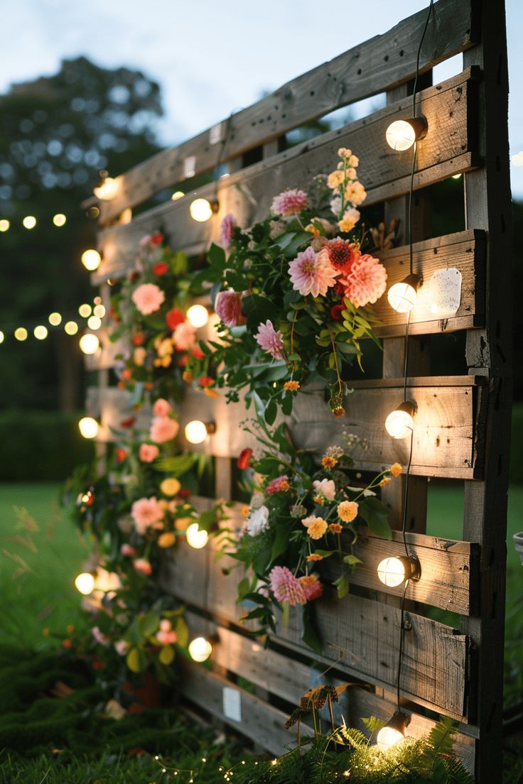 a wooden pallet with flowers and lights on the side is lit up by string lights