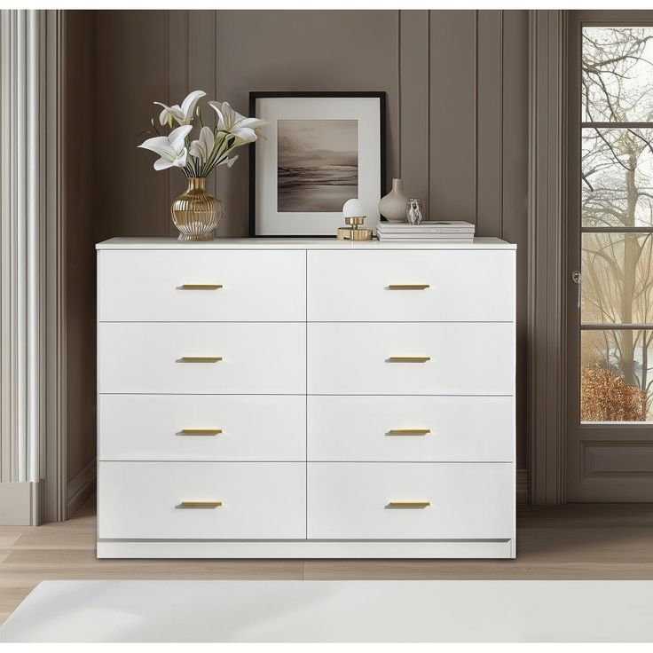 a white dresser with gold handles and drawers in front of a window, framed photograph on the wall