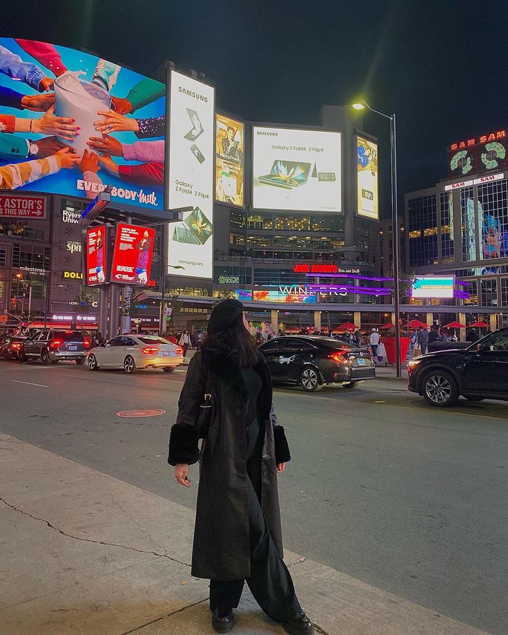 a woman standing on the side of a road in front of tall buildings at night