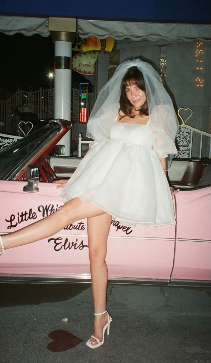 a woman in a short white dress posing next to a pink car