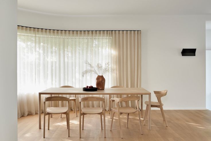 a dining room table and chairs in front of a window with sheered drapes