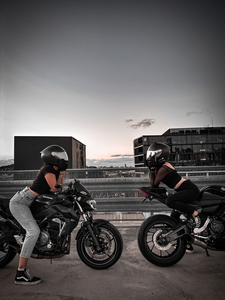 two women sitting on motorcycles in front of a cityscape with buildings behind them