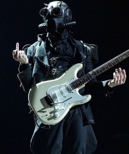 a man in a gas mask holding a guitar and wearing a black outfit with his mouth open