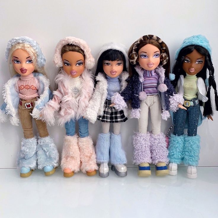 five dolls are lined up against a white wall, one is wearing winter clothes and the other has fur