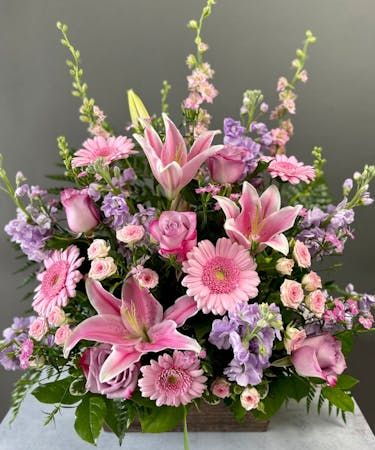 a bouquet of pink and purple flowers in a square vase on a table with greenery