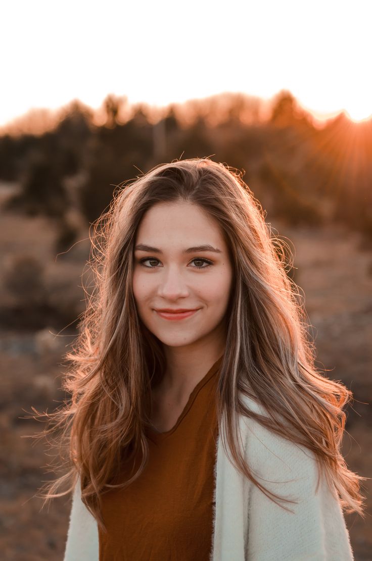 a woman with long hair standing in the middle of a field at sunset, looking into the camera