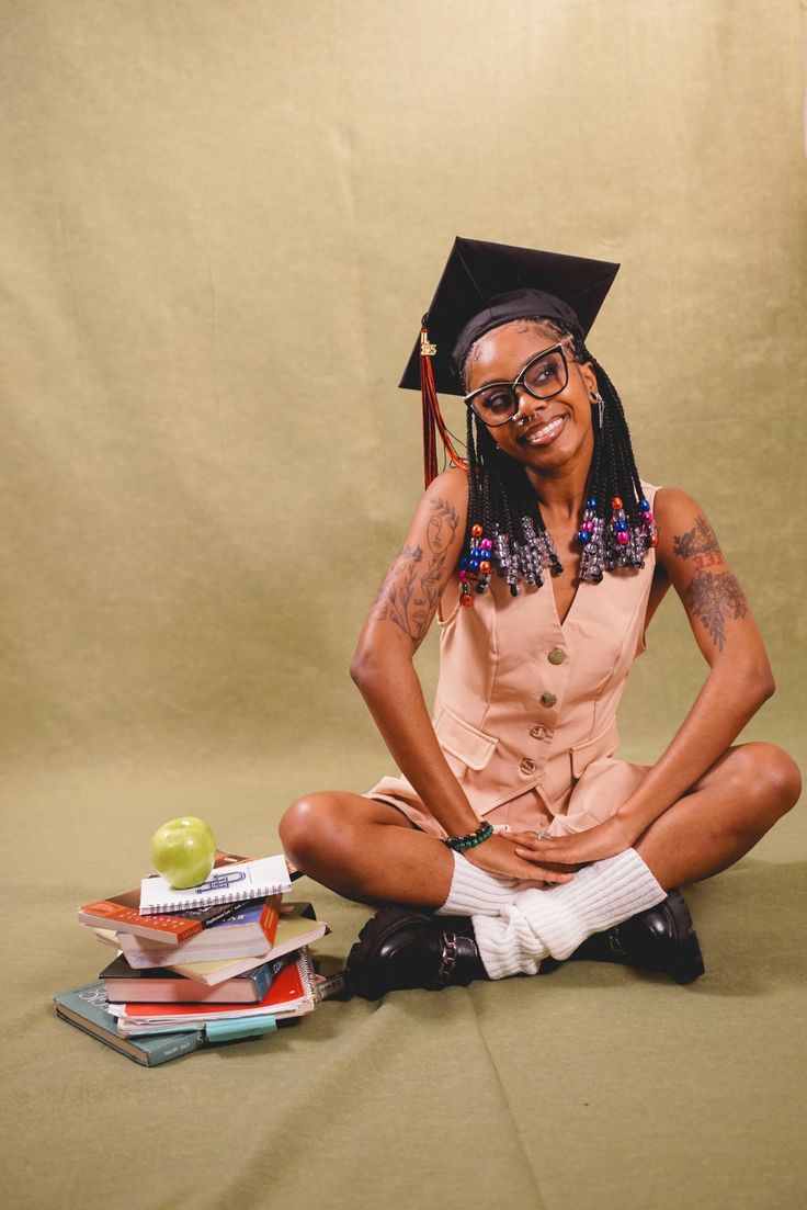 a woman sitting on the ground with books and an apple in front of her, wearing a graduation cap