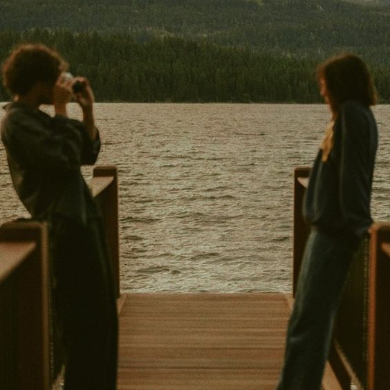 two people standing on a dock looking at the water and mountains in the distance, with one person taking a photo