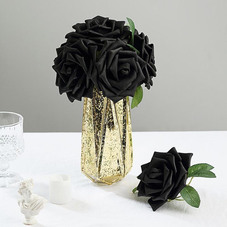 two black roses in a gold vase on a white table cloth next to some candles