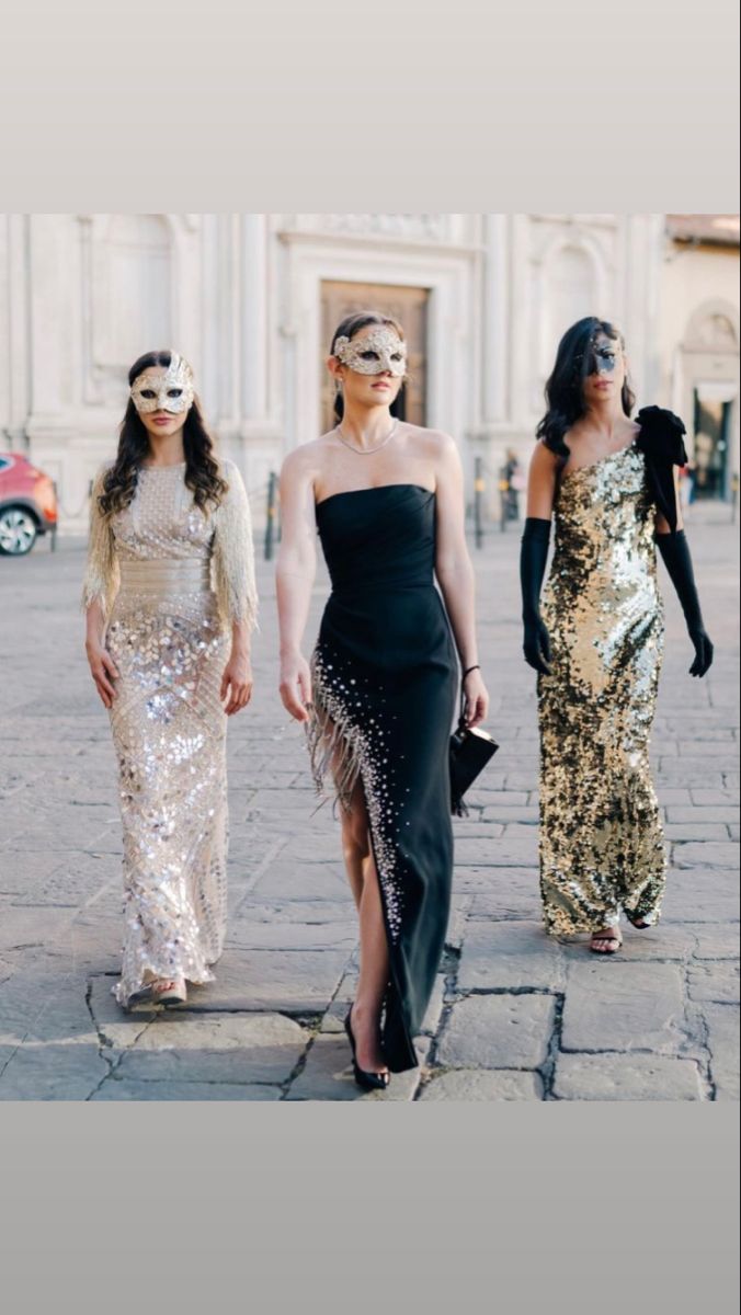 three women in masks walking down the street with one woman wearing a black dress and two are wearing gold dresses