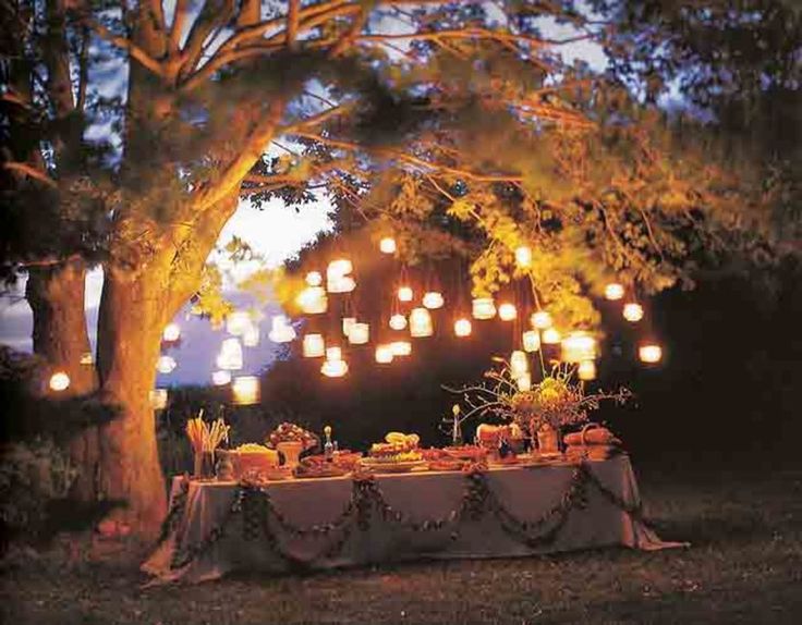 an outdoor dinner table is lit up with lanterns and paper lanterns hanging from the trees